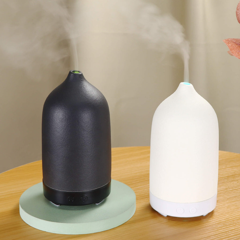 Two ceramic essential oil aroma diffusers, one black and one white, emitting mist. One diffuser is on a wooden table, and the other is on a wooden floor, with a blurred image of a person in the background.