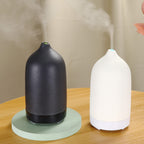 Two ceramic essential oil aroma diffusers, one black and one white, emitting mist. One diffuser is on a wooden table, and the other is on a wooden floor, with a blurred image of a person in the background.